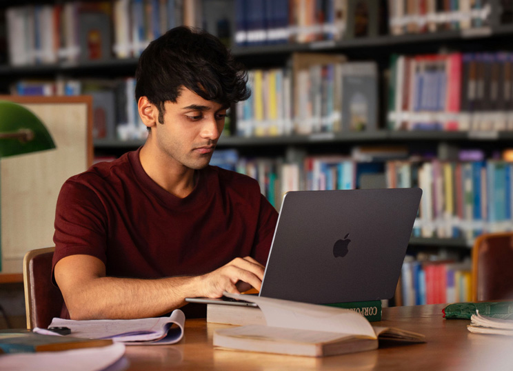 A person working on a MacBook at a library table, surrounded by open books and notes, with bookshelves in the background.